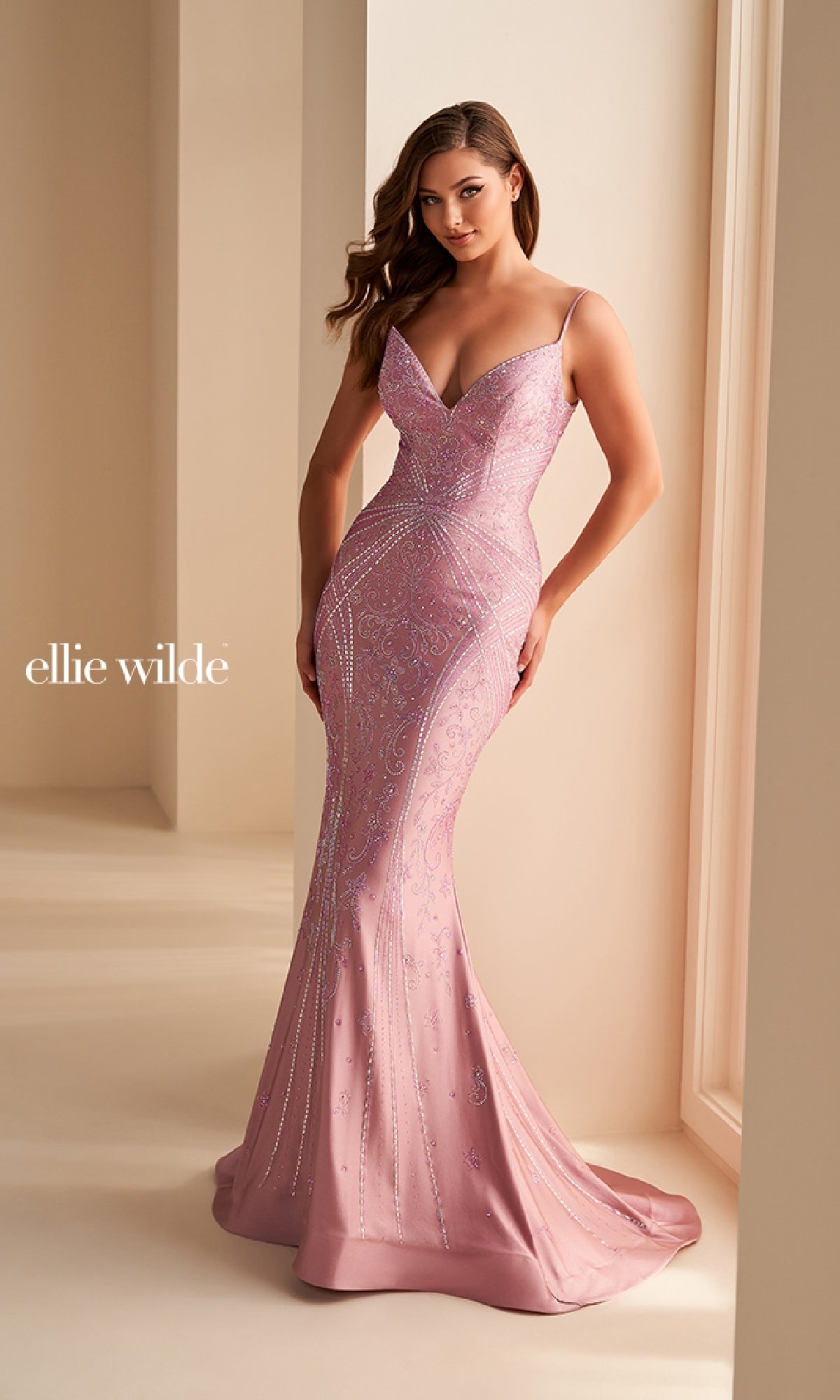 A woman wearing the fitted Ellie Wilde Long Designer Prom Dress EW35002 in light pink, featuring thin straps and shimmering beaded details, stands in a softly lit, neutral-colored room as